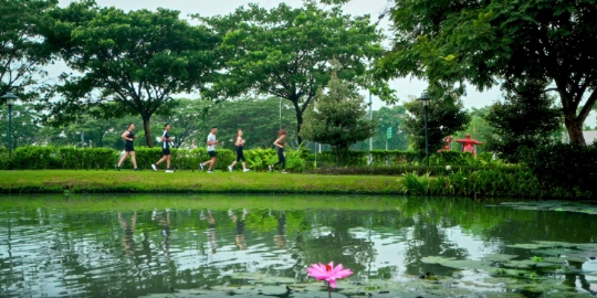 Group of people jogging along a park path beside a pond, with trees and a pink lotus on the water in the foreground.
