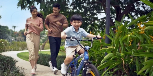 A young boy rides a blue bicycle on a park path as his parents jog behind him among trees and plants in the sunshine.